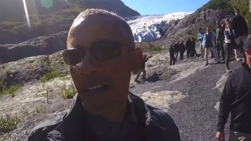 Barack Obama, wearing sunglasses, takes a selfie in front of a glacier in Alaska. Group of people in the background.